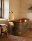 A freestanding copper bathtub with a hammered finish and a matching tall gooseneck faucet, styled in a rustic bathroom with terracotta tile floors, a wooden bench, and a window.