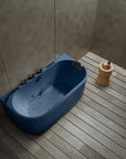 A top view of the Bellastone Doria spa bathtub in Prussian blue with black tap ware, in a modern bathroom with wooden floor and gray walls.